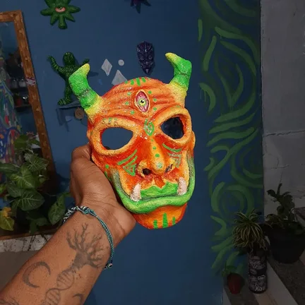 Máscara laranja e verde com chifres, segurada pela mão do artista